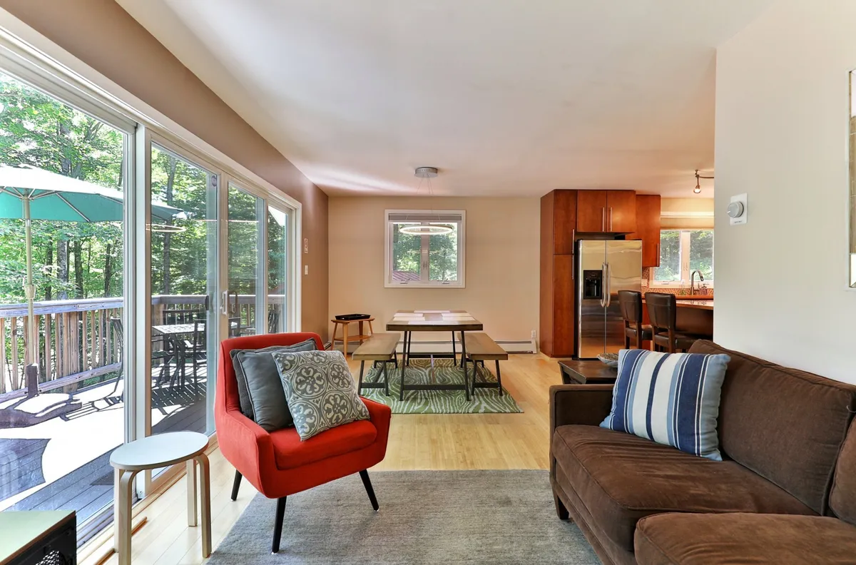 This image captures a warm, welcoming living area that opens to a kitchen and dining space, featuring a brown sofa, red accent chair, and large windows letting in natural light. The spaciousness and cozy arrangement hint at a comfortable base for relaxation after outdoor adventures.
