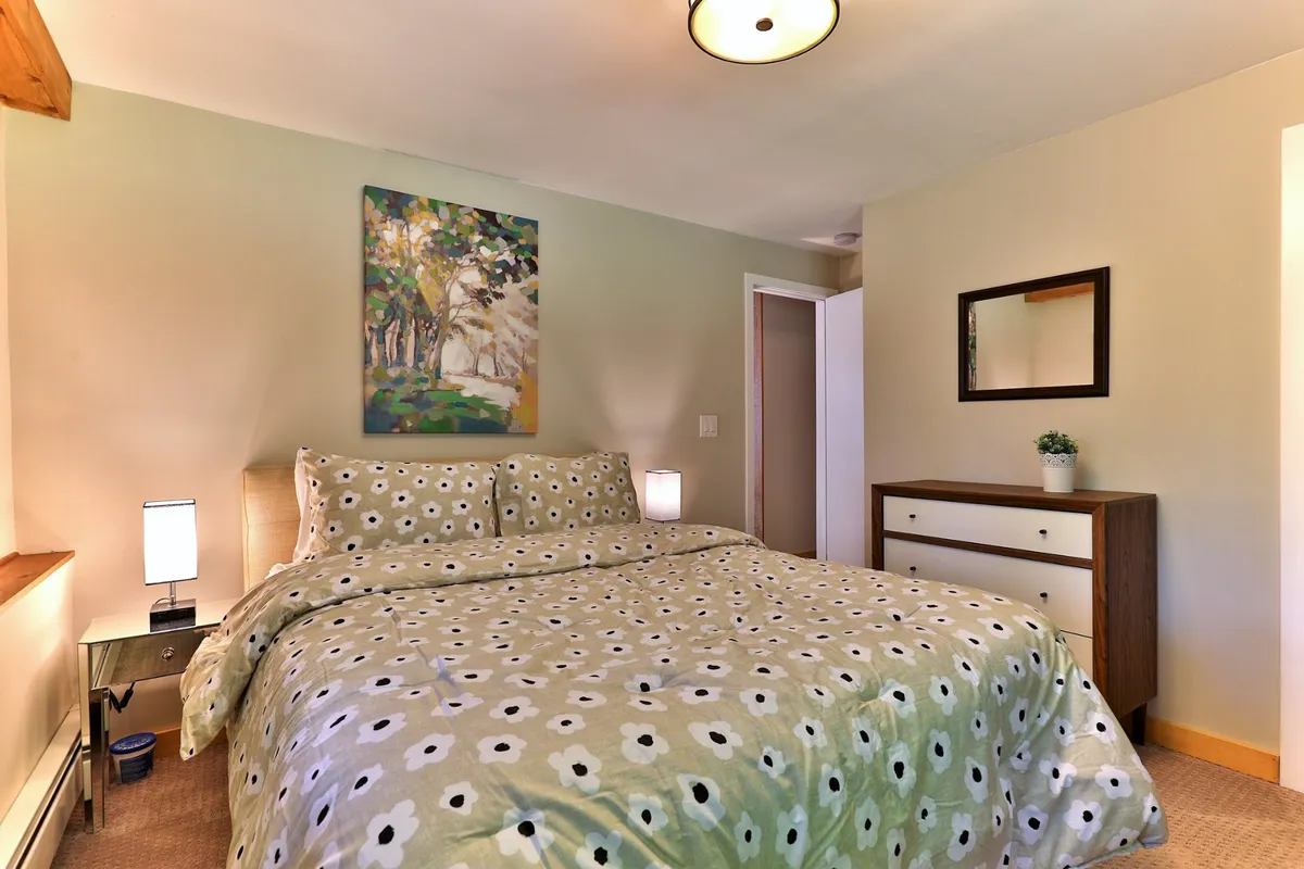 A tranquil bedroom featuring a bed with patterned bedding, colorful artwork on the wall, and a mirrored dresser. Sunlight streams in, creating a restful and inviting atmosphere ideal for unwinding after a day outdoors.