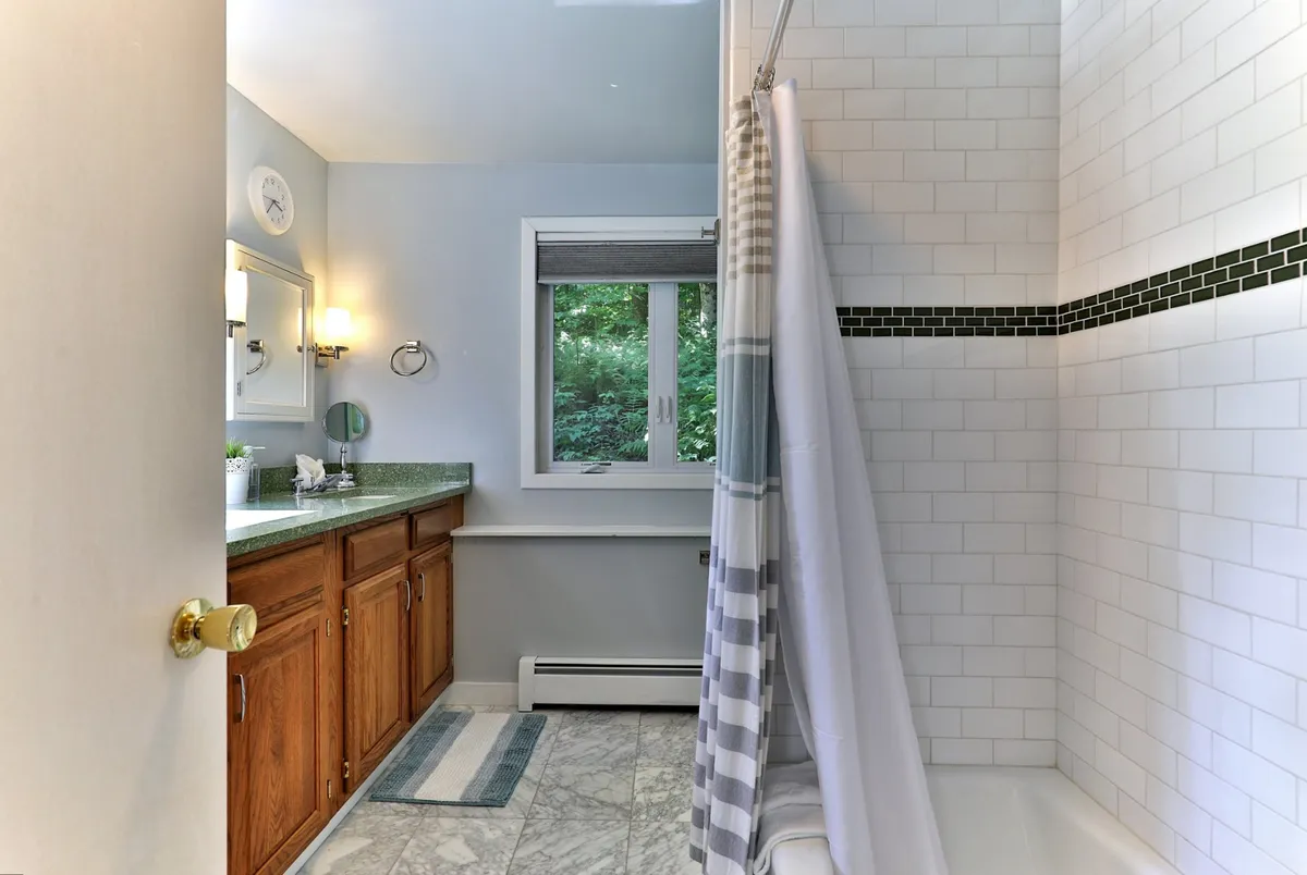 A modern bathroom with a vanity and sink, a window looking out to greenery, and a bathtub with a shower curtain. The space is tranquil and inviting, providing a sense of relaxation and convenience.