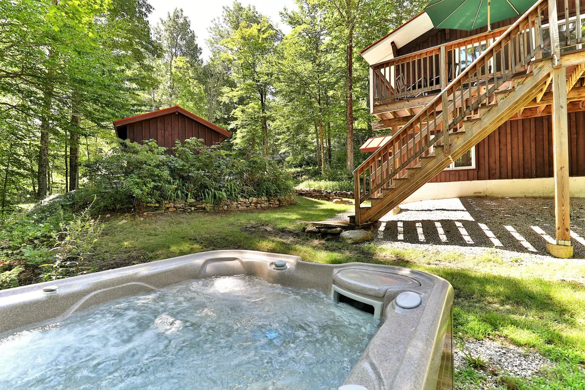 A tranquil wooden deck nestled among Vermont trees, featuring a bubbling hot tub with room for several guests. The cabin is visible in the background, and the deck is bordered by natural greenery, offering a sense of seclusion and privacy. The scene hints at stargazing, relaxation, and time spent in nature.