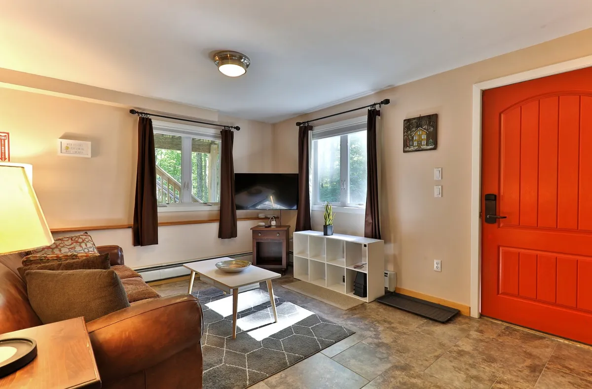 A bright, cheerful living space with an orange front door, a comfy sofa, coffee table, smart TV, and a window letting in natural light. The room is arranged for both relaxation and casual socializing, with the playful door color hinting at a pet-friendly, welcoming atmosphere.