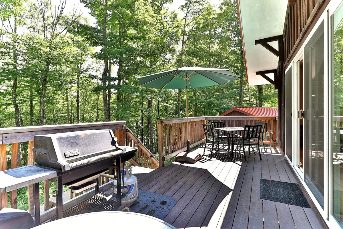 An inviting outdoor dining area on the deck, complete with a grill, dining table under a large umbrella, and wooden railings overlooking the woods. The scene suggests casual al fresco dining, barbecue gatherings, and peaceful moments enjoying Vermont’s outdoors.