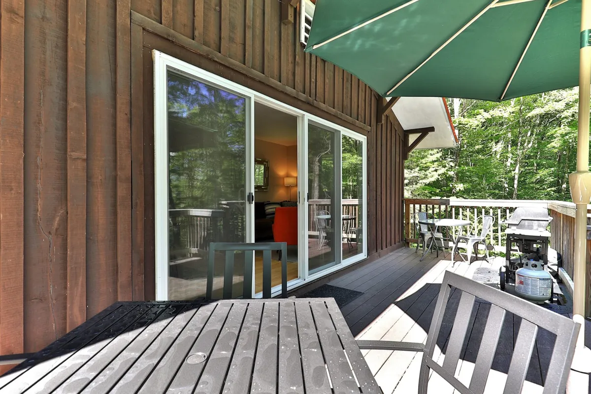 An inviting outdoor wooden deck furnished with a green umbrella, visible through sliding glass doors. The deck is set up for outdoor dining or relaxing, surrounded by greenery that hints at the natural Vermont setting. The space feels private yet open, ideal for gatherings or unwinding after a day out.