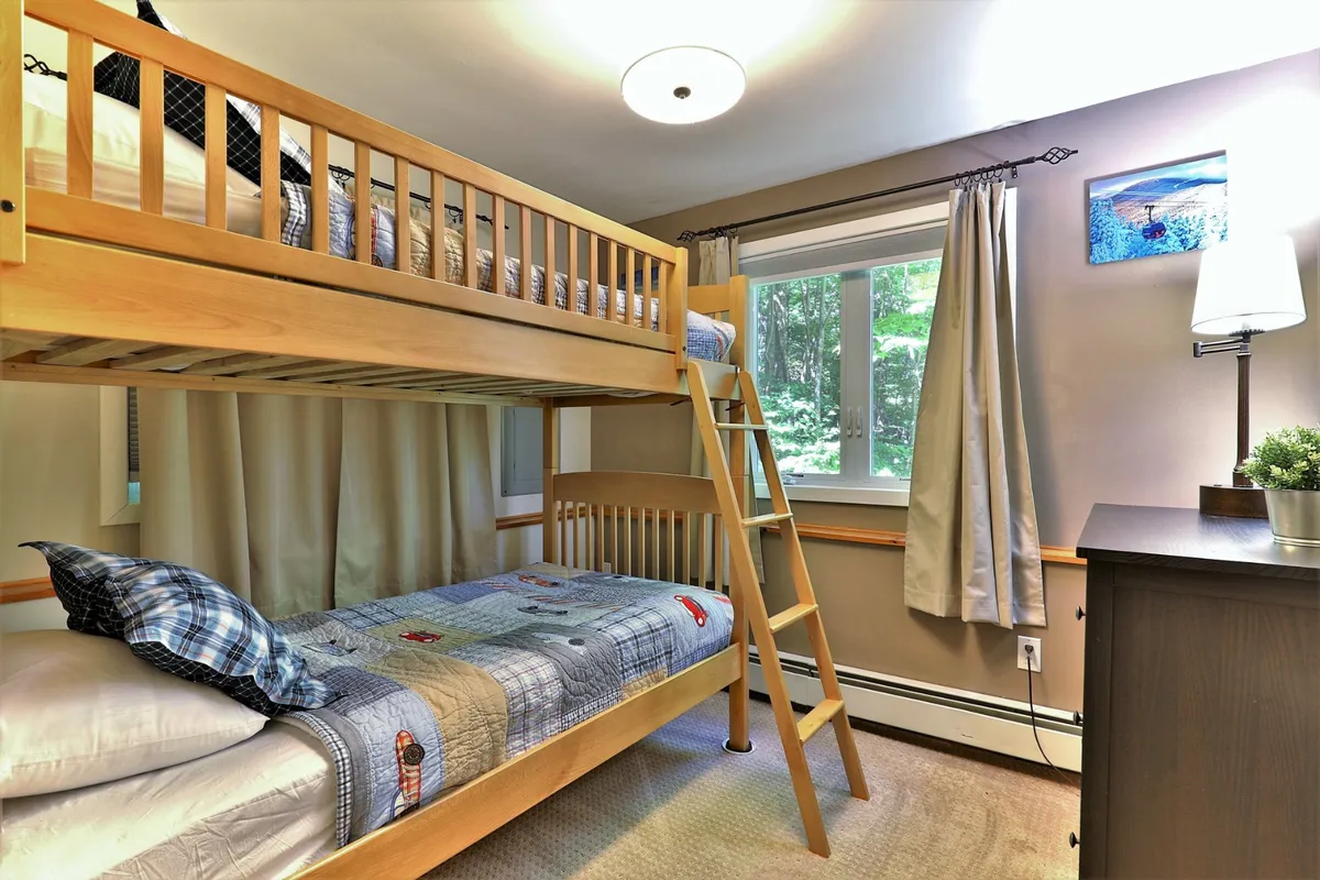 An interior shot of a cozy bunk bed setup next to a window, complete with a nightstand. The scene evokes warmth and playful comfort—ideal for children after a long day on the slopes.