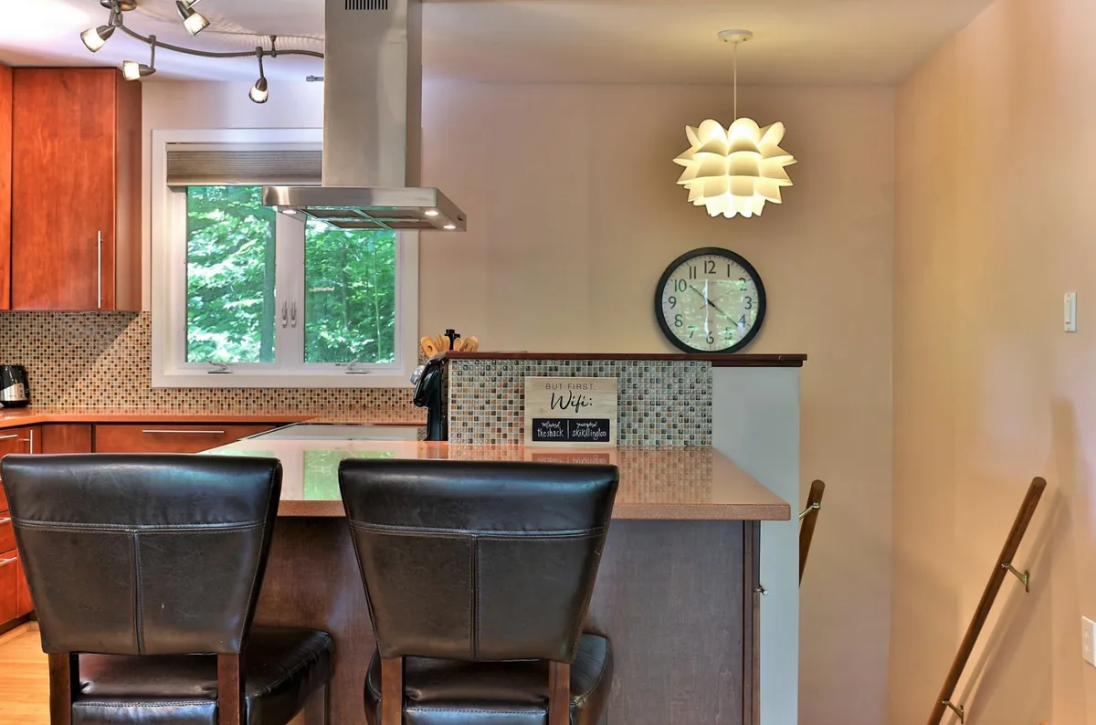 The kitchen features a modern island with bar stools, pendant lighting, a wall clock, and a window view, creating a social hub for casual dining and morning routines.