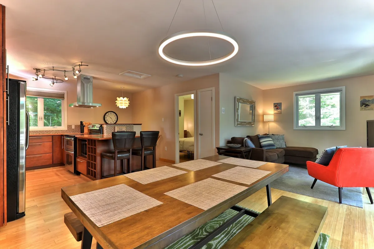 The image showcases a cozy open-concept living and dining space, featuring a large dining table perfect for group meals or game nights, a modern kitchen area, a plush sofa, and a striking red accent chair—all arranged to foster togetherness and relaxation.