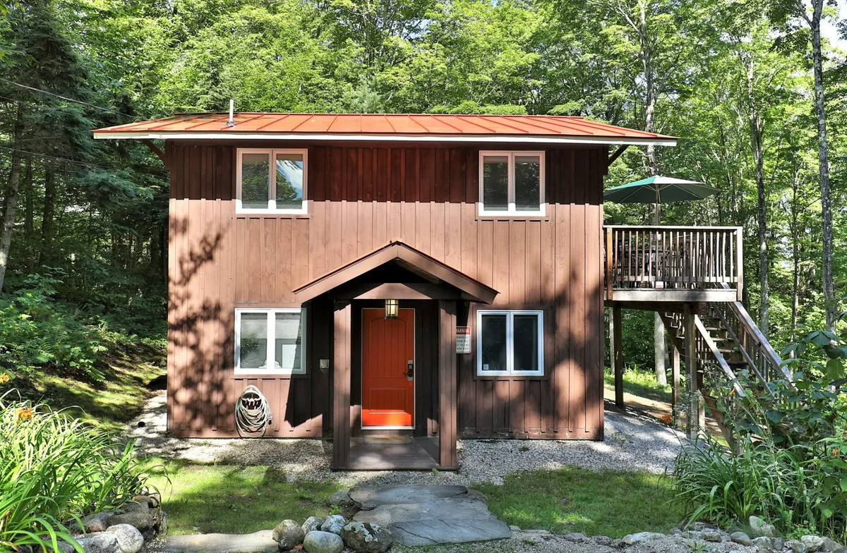 The cabin’s welcoming wooden exterior with a bright orange door, a deck furnished with an umbrella, surrounded by lush greenery. The ambiance is tranquil and inviting, perfect for relaxing, hosting friends, or simply soaking in the scenery.